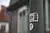Nahaufnahme eines weißen Aufklebers mit Barcode auf einem grauen Pfosten, im Hintergrund unscharfes Wohnhaus mit grünen Fensterläden.